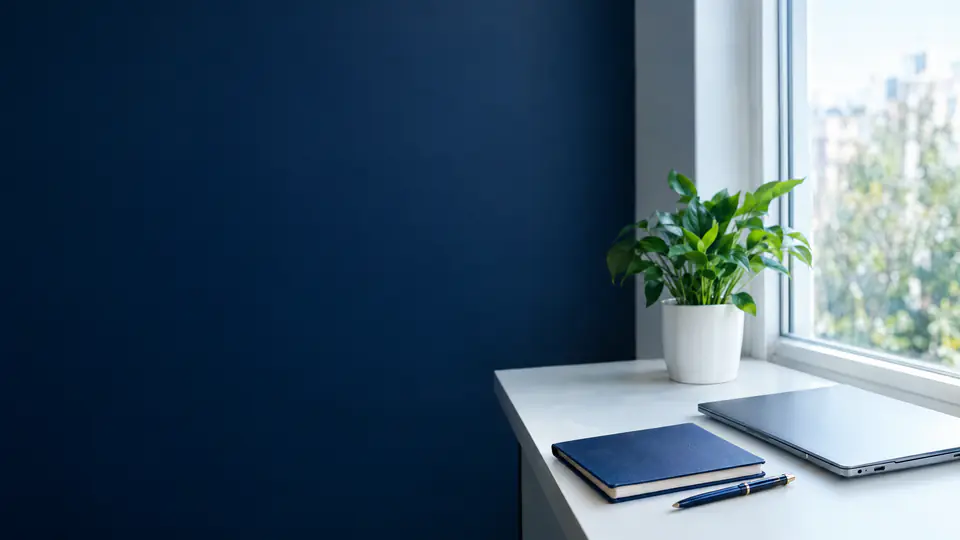 Desk by a bright window with a navy notebook, a pen, a closed laptop and a plant, against a navy wall