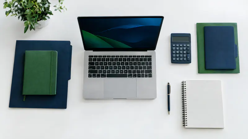Overhead view of a laptop, calculator, notebook and navy and green folders on a white desk