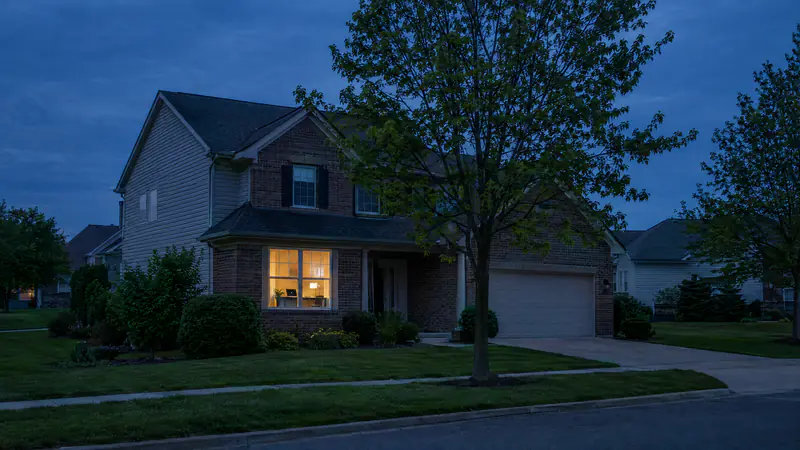 Quiet residential street at dusk with a home office window glowing and a laptop on the desk